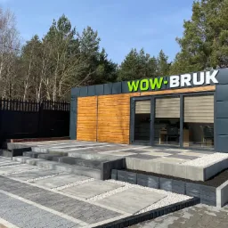 Ekspozycja różnego rodzaju nawierzchni z kostki brukowej przed biurem firmy WOW-BRUK, z widocznym logo na budynku i czarnym ogrodzeniem w tle.