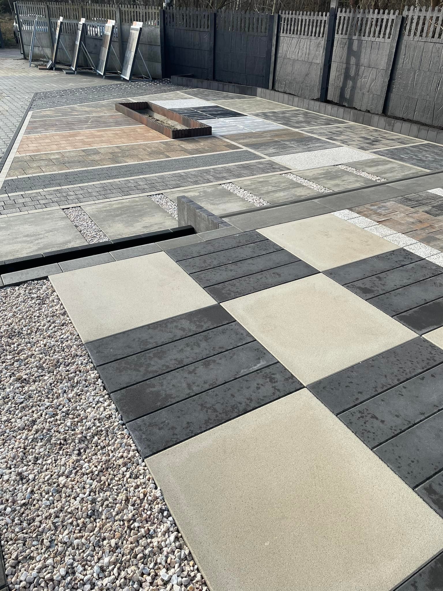Ekspozycja różnych wzorów i kolorów nawierzchni z kostki, w tym szachownica z szarych i beżowych prostokątnych elementów, oraz fragmenty ogrodzeń betonowych w tle.