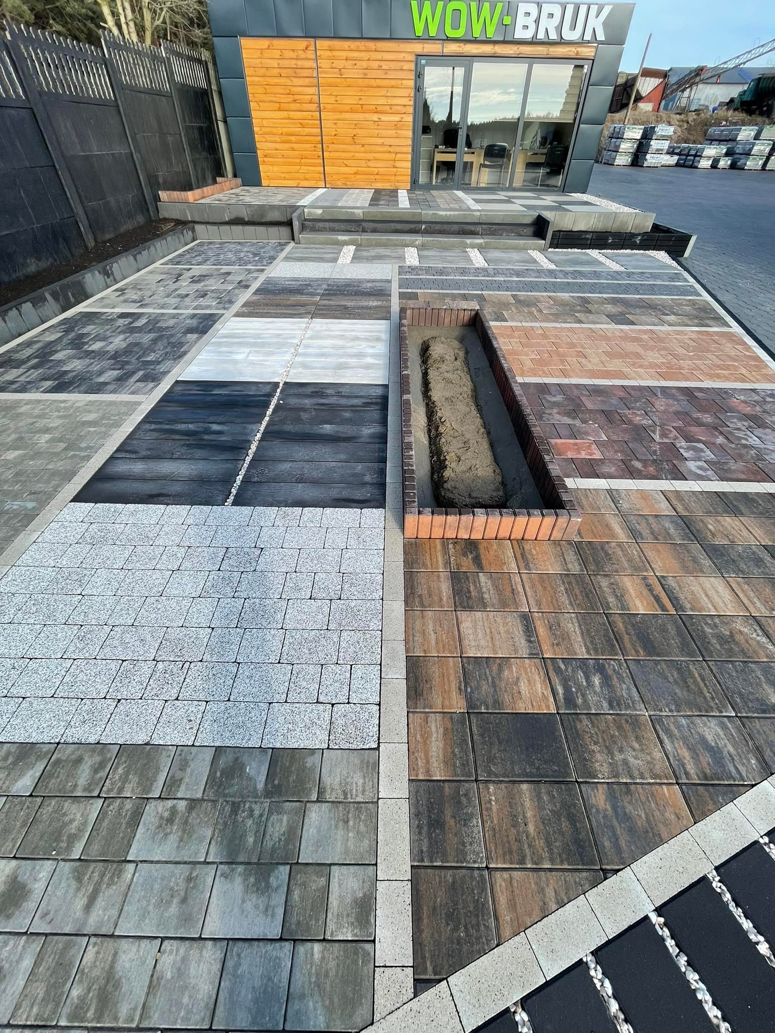 Ekspozycja różnych wzorów i kolorów nawierzchni ułożonych obok siebie, w tym fragmentu z ziemią w obramowaniu z cegieł, przed budynkiem z logo 'WOW-BRUK'.