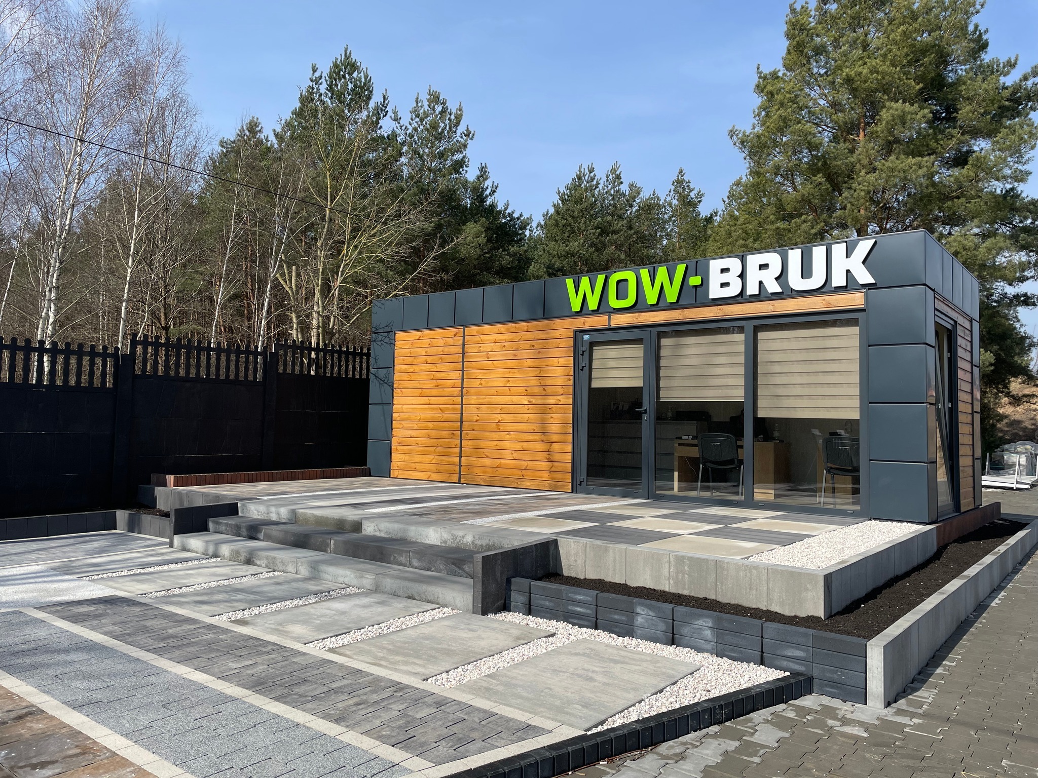 Ekspozycja różnego rodzaju nawierzchni z kostki brukowej przed biurem firmy WOW-BRUK, z widocznym logo na budynku i czarnym ogrodzeniem w tle.