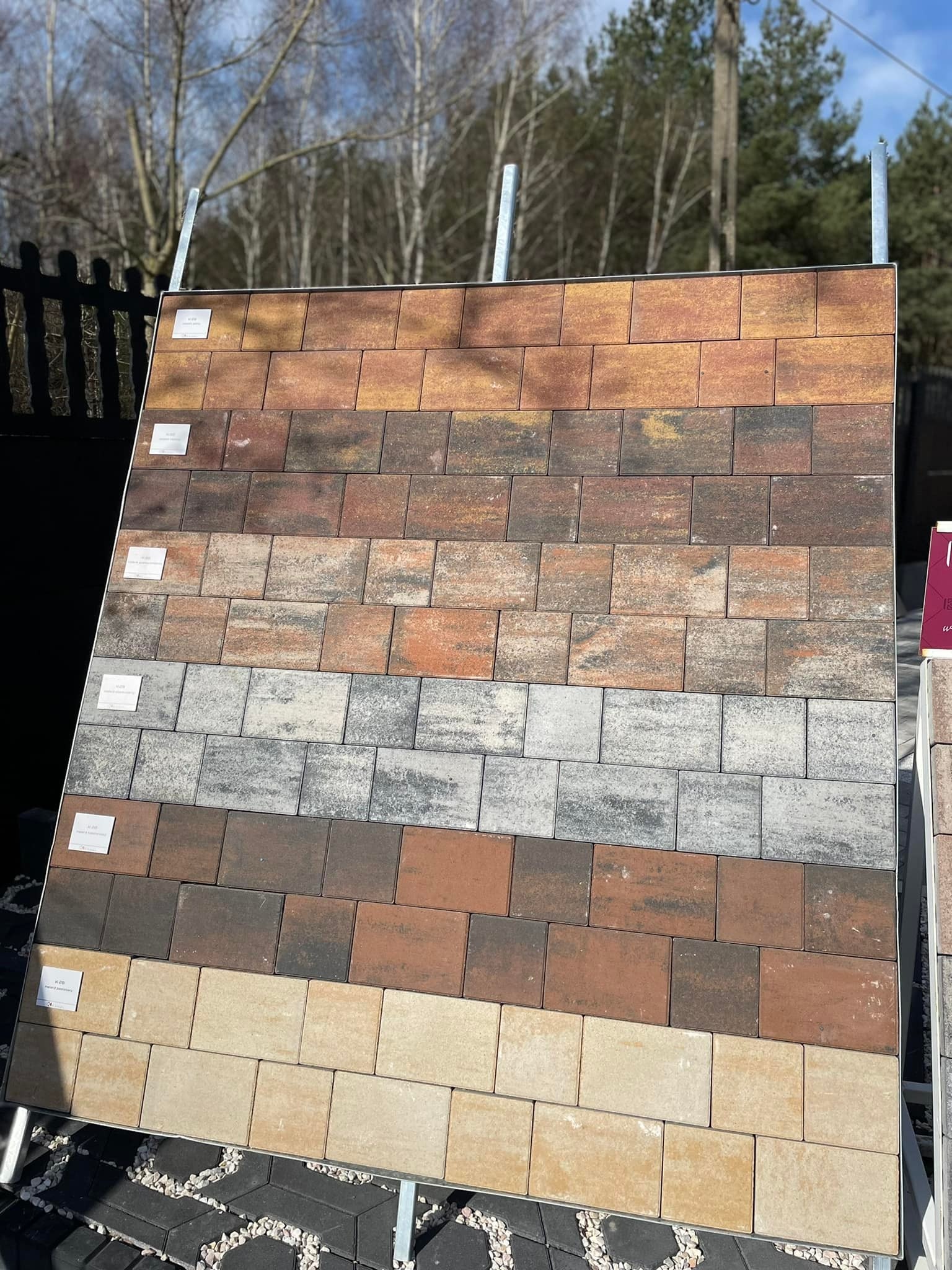 Ekspozycja różnych kolorów i wzorów kostki betonowej na stojaku, widoczny fragment ogrodzenia i drzewa w tle.