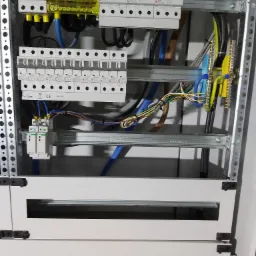 Rozdzielnica elektryczna z zainstalowanymi bezpiecznikami, zabezpieczeniami przepięciowymi i okablowaniem, widoczne szyny montażowe i elementy metalowej obudowy.