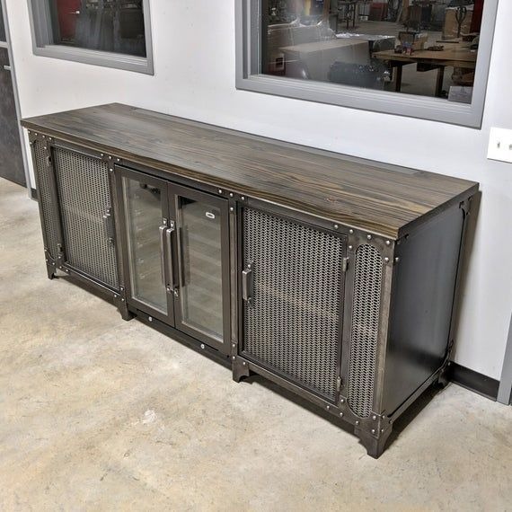 Industrialna szafka RTV z drewnianym blatem, metalową siatką i szklanymi drzwiczkami na tle betonowej podłogi i okna warsztatu.