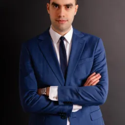 Portret mężczyzny w niebieskim garniturze, białej koszuli i czarnym krawacie, z zegarkiem na ręce i splecionymi rękami na tle ciemnoszarego tła.