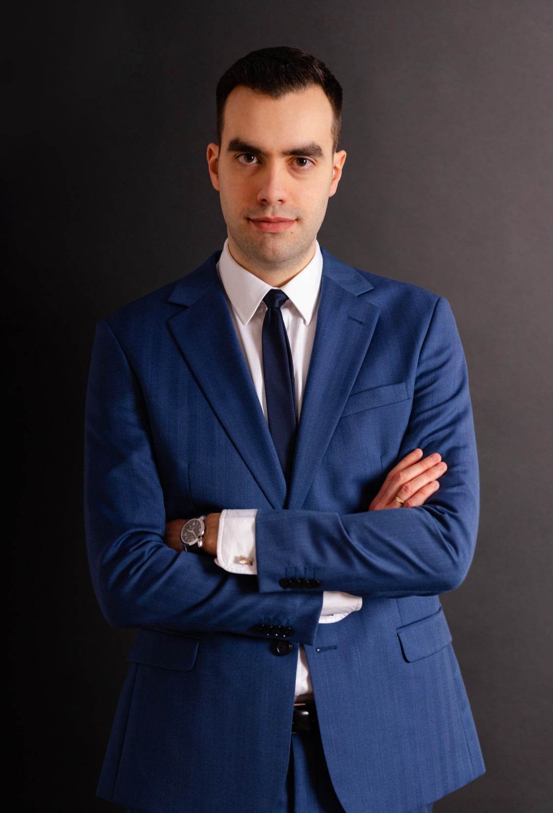 Portret mężczyzny w niebieskim garniturze, białej koszuli i czarnym krawacie, z zegarkiem na ręce i splecionymi rękami na tle ciemnoszarego tła.