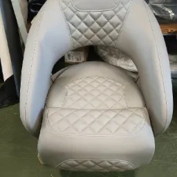 Jasnoszare, pikowane siedzisko z oparciem w kształcie litery U, widok z przodu na tle materiałów i kartonów.