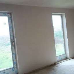 Surowe, jasne wnętrze z dwoma oknami z profilami VEKA po nałożeniu świeżych, szarych tynków maszynowych na ścianach i suficie, widoczne niedoskonałości i nierówności na powierzchni.