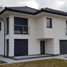 Nowoczesny dom jednorodzinny z grafitowymi roletami zewnętrznymi, czarnymi oknami i bramą garażową, białą elewacją i ciemną dachówką, otoczony trawnikiem i ozdobnymi nasadzeniami.