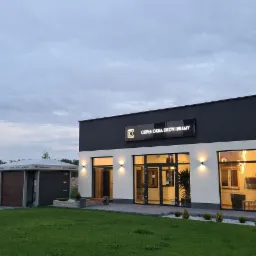 Budynek firmy oferującej okna, drzwi i bramy, z ekspozycją bram garażowych na zewnątrz, widoczne flagi z logo firmy.