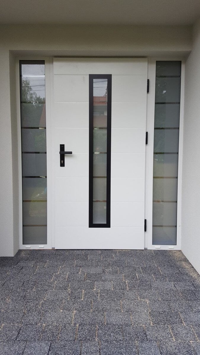 Białe drzwi wejściowe z czarną ramką okienną i bocznymi panelami szklanymi, zamontowane w nowym budynku z szarą kostką brukową przed wejściem.