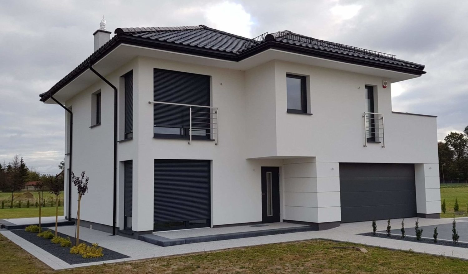 Nowoczesny dom jednorodzinny z grafitowymi roletami zewnętrznymi, czarnymi oknami i bramą garażową, białą elewacją i ciemną dachówką, otoczony trawnikiem i ozdobnymi nasadzeniami.