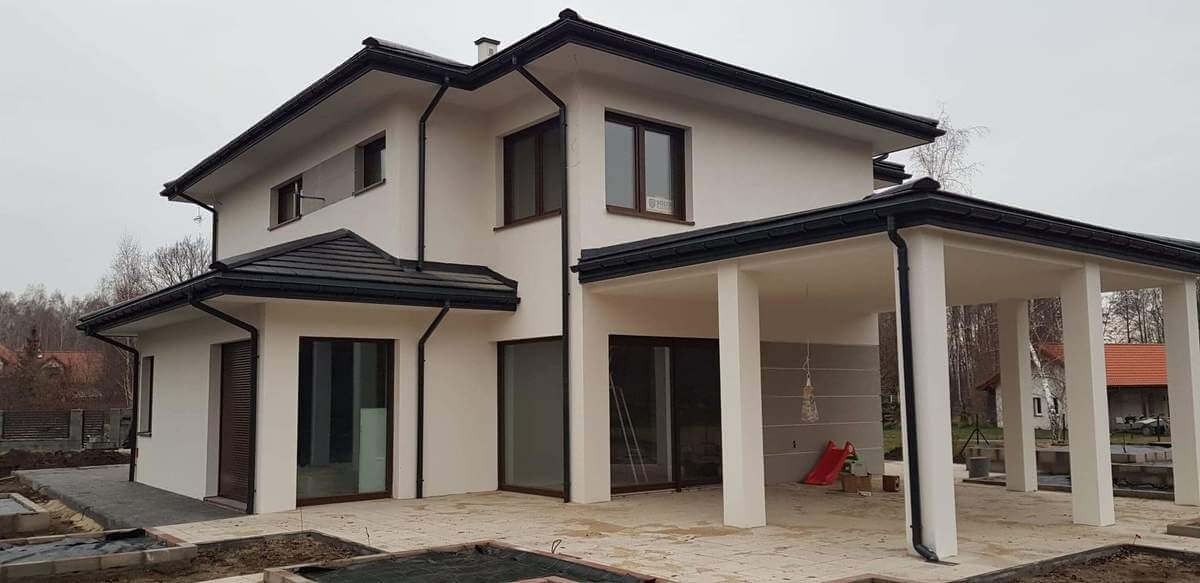 Nowoczesny dom jednorodzinny z białymi ścianami i ciemnobrązowymi oknami PCV oraz zadaszonym tarasem z kolumnami, widok z zewnątrz w pochmurny dzień.