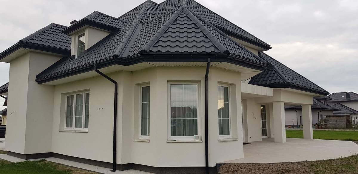 Elewacja nowo wybudowanego domu jednorodzinnego z białymi oknami z szybami podzielonymi na sześć części i ciemną dachówką.
