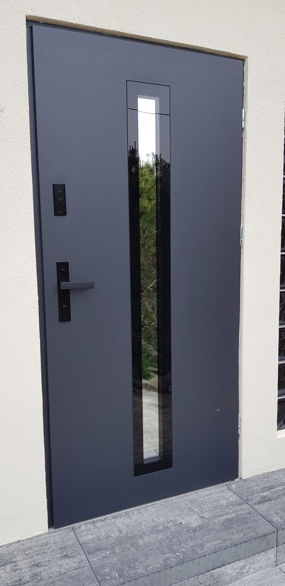 Nowoczesne, grafitowe drzwi wejściowe z wąskim, pionowym przeszkleniem, widoczne z zewnątrz, osadzone w jasnej elewacji budynku.
