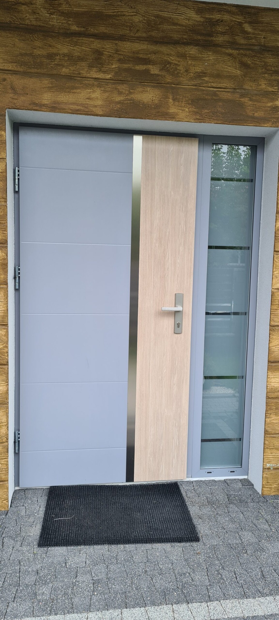 Szare drzwi wejściowe z panelem imitującym drewno i przeszkleniem, widok z zewnątrz, czarna wycieraczka przed drzwiami.