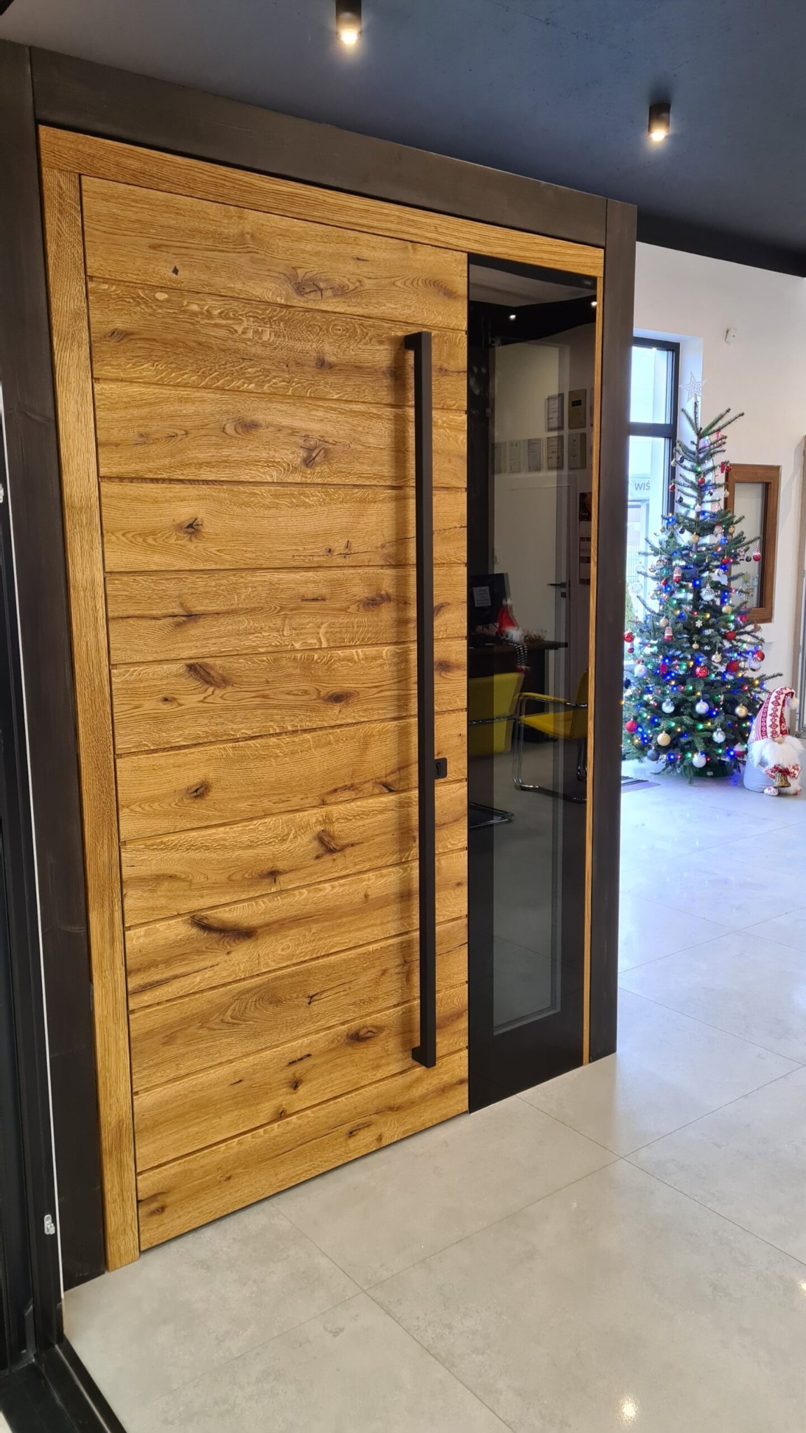 Nowoczesne drzwi wejściowe z drewnianą okładziną i czarną ramą, widoczne przez szybę wnętrze z choinką.