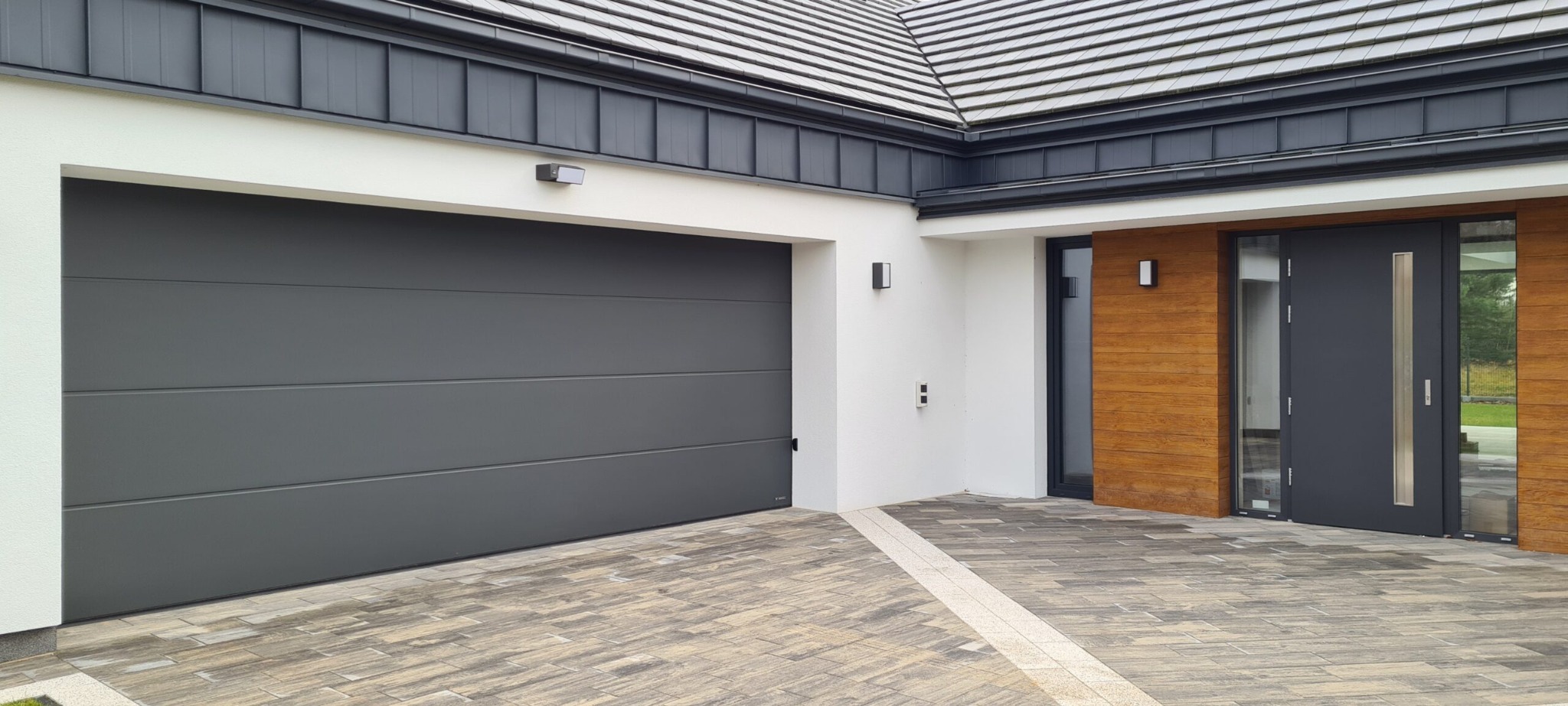 Nowoczesna brama garażowa segmentowa w kolorze grafitowym, z poziomymi przetłoczeniami, zamontowana w białej elewacji domu z drewnianą okładziną przy drzwiach wejściowych i kostką brukową...