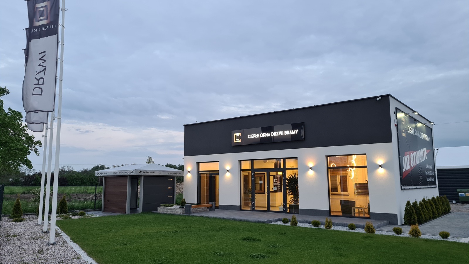 Budynek firmy oferującej okna, drzwi i bramy, z ekspozycją bram garażowych na zewnątrz, widoczne flagi z logo firmy.