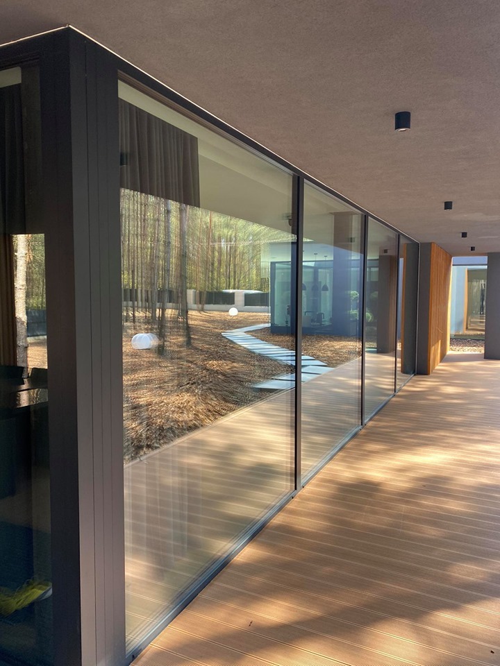 Nowoczesny przeszklony korytarz z widokiem na zalesiony ogród i ścieżkę z białych płyt, oświetloną kulami.