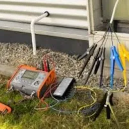 Pomarańczowy miernik elektryczny z podłączonymi przewodami i krokodylkami na trawie przy budynku z białym sidingiem, widoczne drobne kamienie i rura kanalizacyjna.