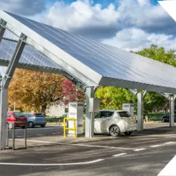 Zadaszony parking z panelami słonecznymi i stanowiskami do ładowania samochodów elektrycznych, zaparkowany biały Nissan Leaf, jesienne drzewa w tle.