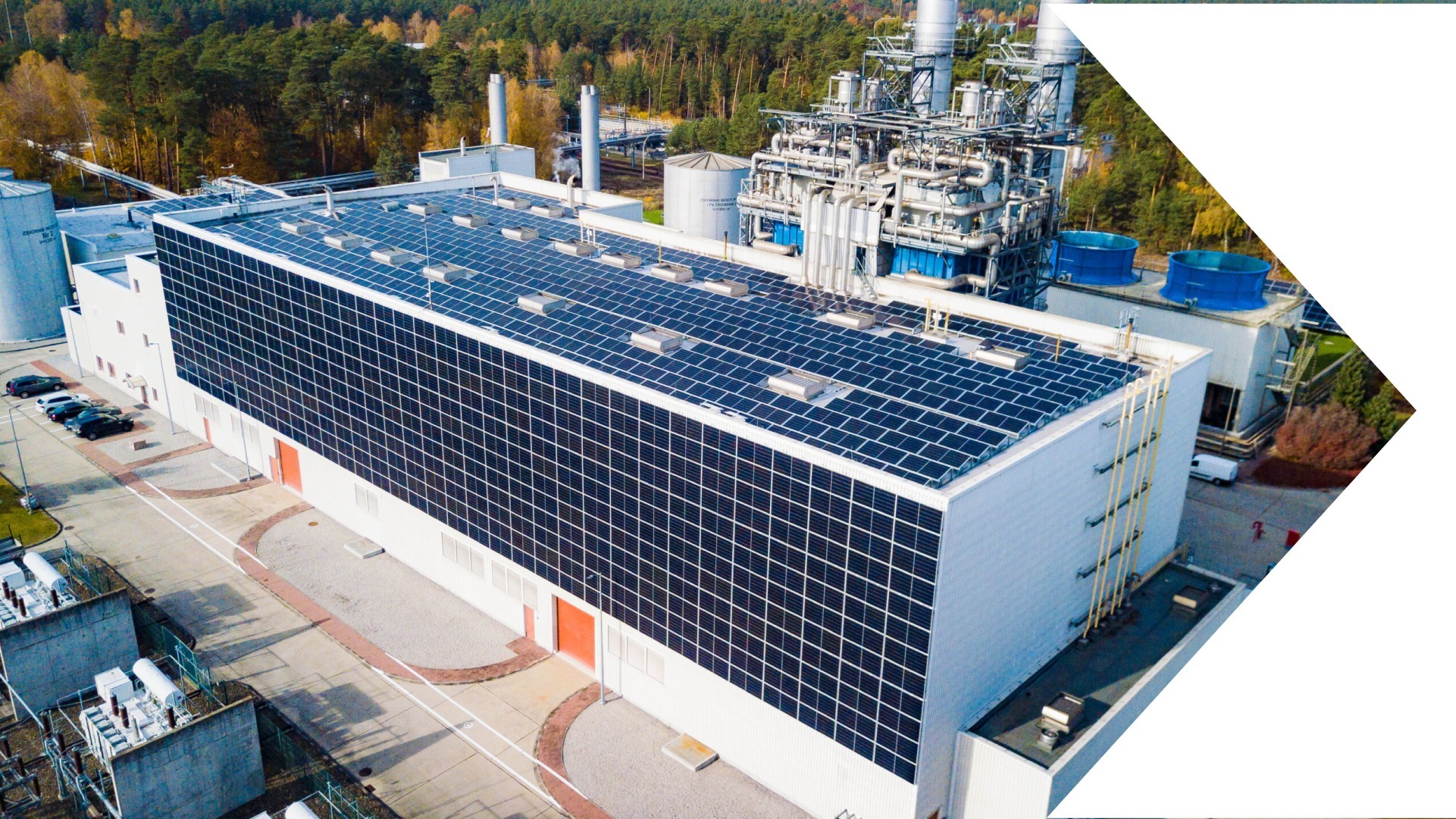 Widok z góry na duży budynek przemysłowy pokryty panelami słonecznymi, z widocznymi elementami infrastruktury energetycznej i otaczającym lasem.