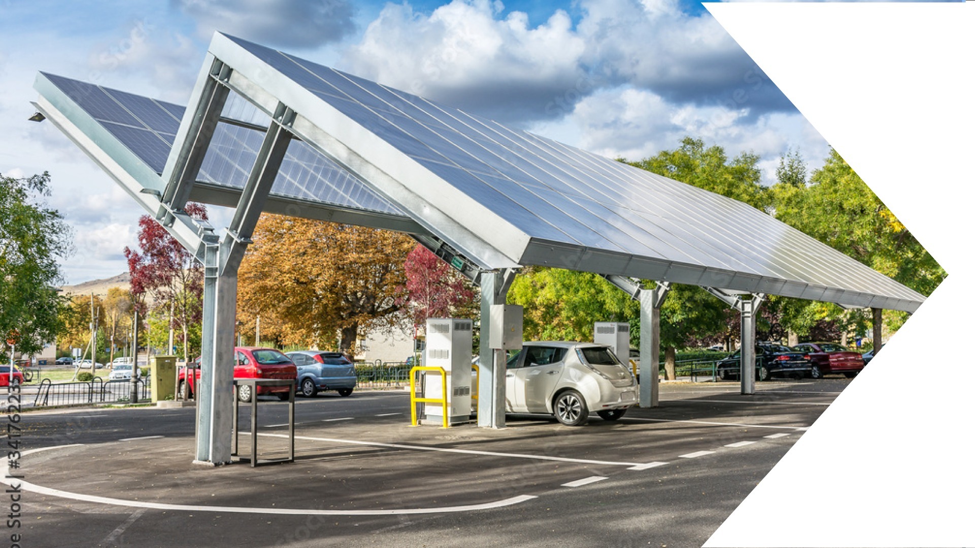 Zadaszony parking z panelami słonecznymi i stanowiskami do ładowania samochodów elektrycznych, zaparkowany biały Nissan Leaf, jesienne drzewa w tle.