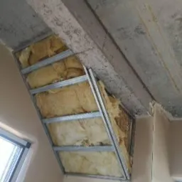 Sufit z zamontowaną metalową konstrukcją pod płyty kartonowo-gipsowe wypełnioną wełną mineralną, widoczne okno dachowe i surowy betonowy strop.