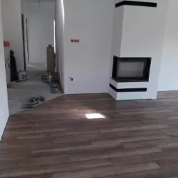 Nowoczesny salon w trakcie wykończenia: ciemna podłoga imitująca drewno, białe ściany z kominkiem obudowanym na biało z czarnymi akcentami, widoczne ślady prac remontowych w przejściu do kolejnego...