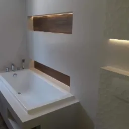 Minimalistyczna łazienka w Luszowicach z białą wanną obudowaną płytami, podświetlona wnęka z drewnopodobnymi panelami i dekoracyjna ściana z tr&oacute;jwymiarową fakturą.