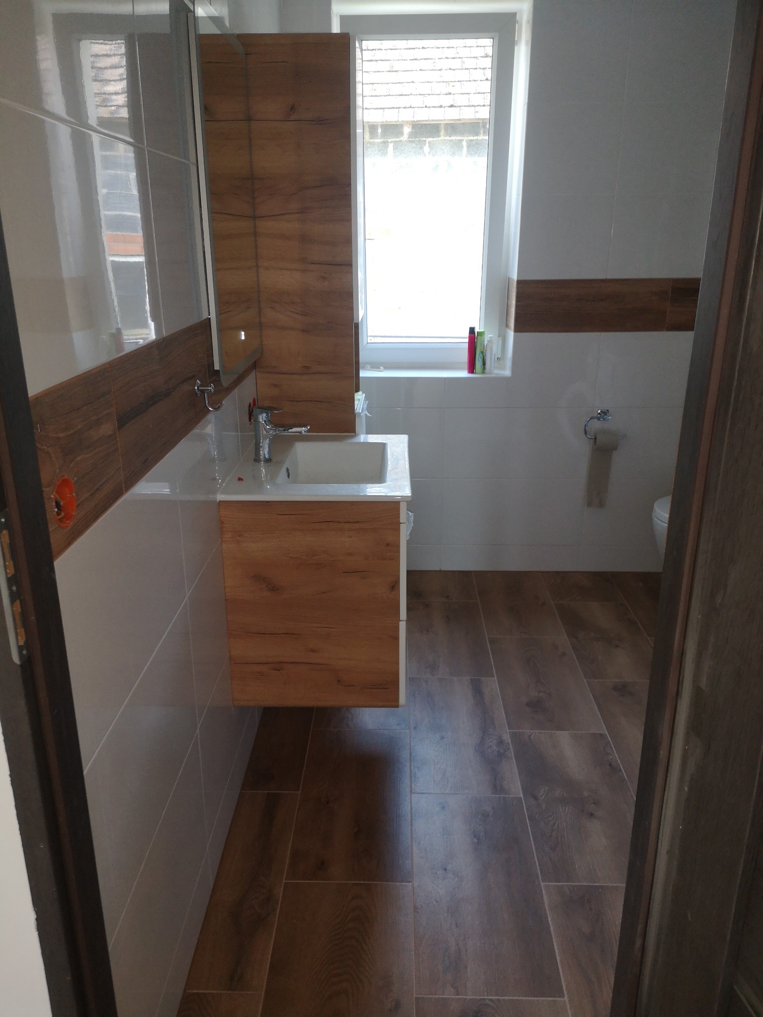 Nowoczesna łazienka z drewnopodobnymi płytkami na podłodze i ścianie, wisząca szafka pod umywalką z lustrem, widoczne gniazdko elektryczne w ścianie i fragment toalety.