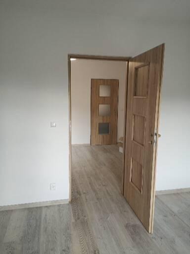 Otwarta, drewniana, wewnętrzna drzwi z trzema kwadratowymi okienkami, widok na sąsiednie pomieszczenie z identycznymi drzwiami, jasna podłoga imitująca drewno, białe ściany, widoczne gniazdko...