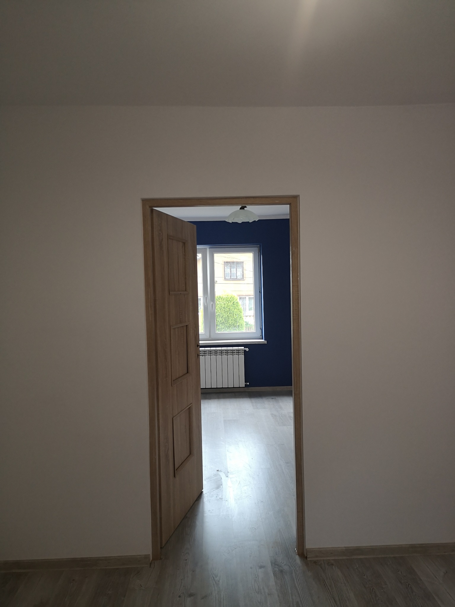 Otwarta drewniana trzyskrzydłowa drzwi wewnętrzne w nowo wyremontowanym pokoju z pomalowaną na granatowo ścianą, białym oknem i grzejnikiem, podłoga wyłożona panelami.