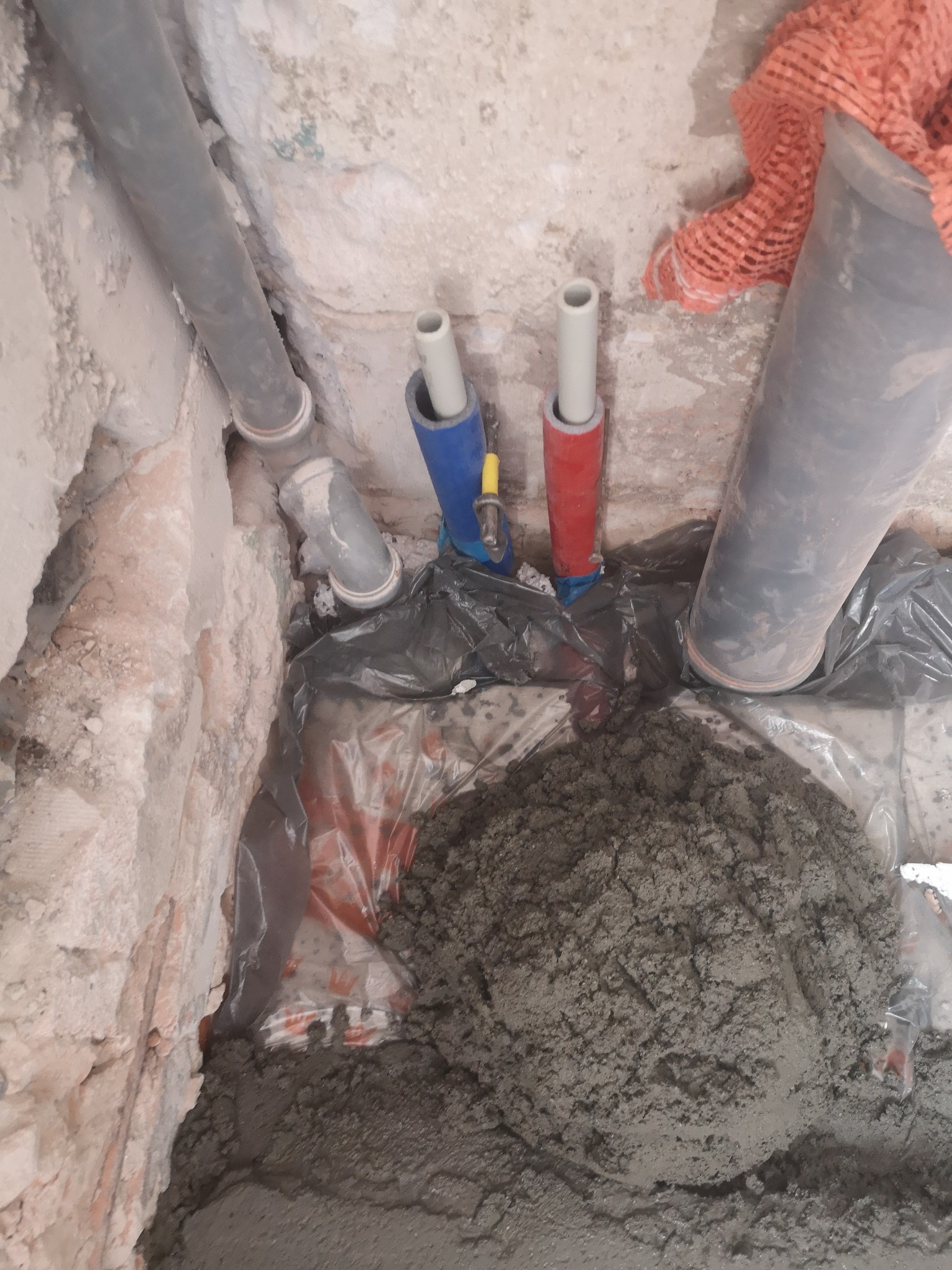 Przygotowanie podłoża pod posadzkę w łazience: widoczne rury instalacji wodnej z niebieskimi i czerwonymi złączkami, szara rura kanalizacyjna, folia ochronna i przygotowana zaprawa cementowa.