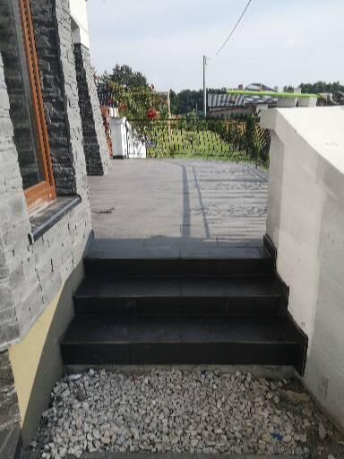 Czarne, płytkowe schody zewnętrzne prowadzące na taras, z widocznym fragmentem elewacji budynku wykończonej kamieniem dekoracyjnym i podsypką z drobnych kamieni u podstawy schodów.