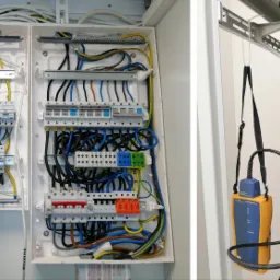 Otwarta skrzynka elektryczna z bezpiecznikami i okablowaniem oraz tester sieci komputerowej wiszący obok panelu krosowniczego.