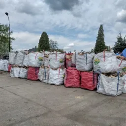 Stos worków z pociętym drewnem ułożonych na betonowym placu, widoczne drzewa w tle i pochmurne niebo.