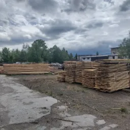 Stosy świeżo przetartego drewna ułożone na placu tartaku, widoczne napisy cen na niektórych pakietach, w tle budynek i pochmurne niebo.