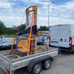 FS-Lechowicz Krzysztof Lechowicz - Nowy wózek widłowy Jungheinrich, owinięty folią i zabezpieczony pomarańczowymi pasami transportowymi, umieszczony na przyczepie samochodowej, gotowy do transportu, z widocznym fragmentem białego...