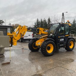 FS-Lechowicz Krzysztof Lechowicz - Żółto-czarny wózek widłowy teleskopowy JCB podnosi betonowy element konstrukcyjny na mokrym placu budowy, w tle budynek i zaparkowany biały samochód dostawczy.