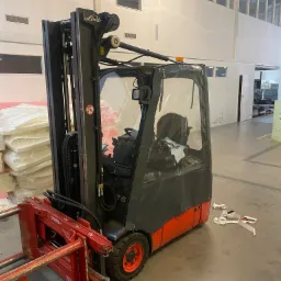 Wózek widłowy Linde E15 z czerwonym osprzętem Bolzoni Auramo do transportu materiałów w magazynie, widoczne opakowania z pianką i białe taśmy na podłodze.