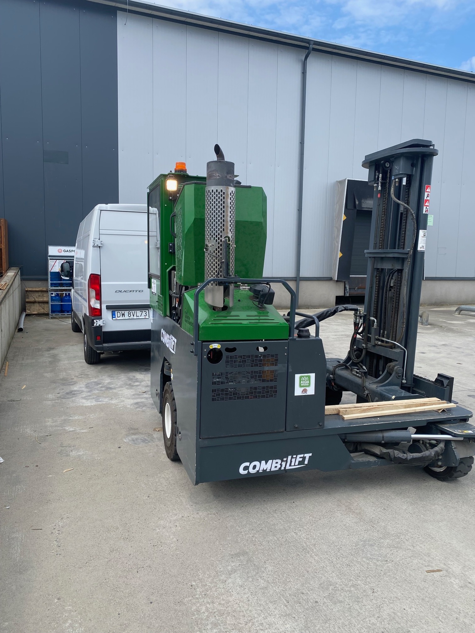 Wielokierunkowy wózek widłowy Combilift z zielonym korpusem i podniesionym masztem, zaparkowany na betonowym placu przed szarą halą, obok białego dostawczego Ducato.