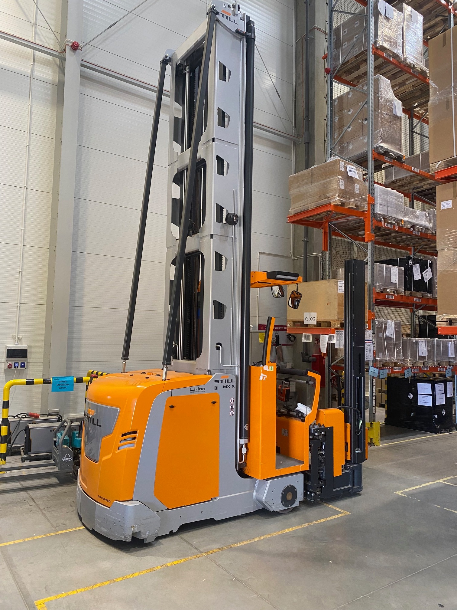 Pomarańczowo-szary wózek widłowy wysokiego składowania marki Still, model MX-X z akumulatorem Li-Ion, stojący w magazynie z regałami wypełnionymi kartonami.
