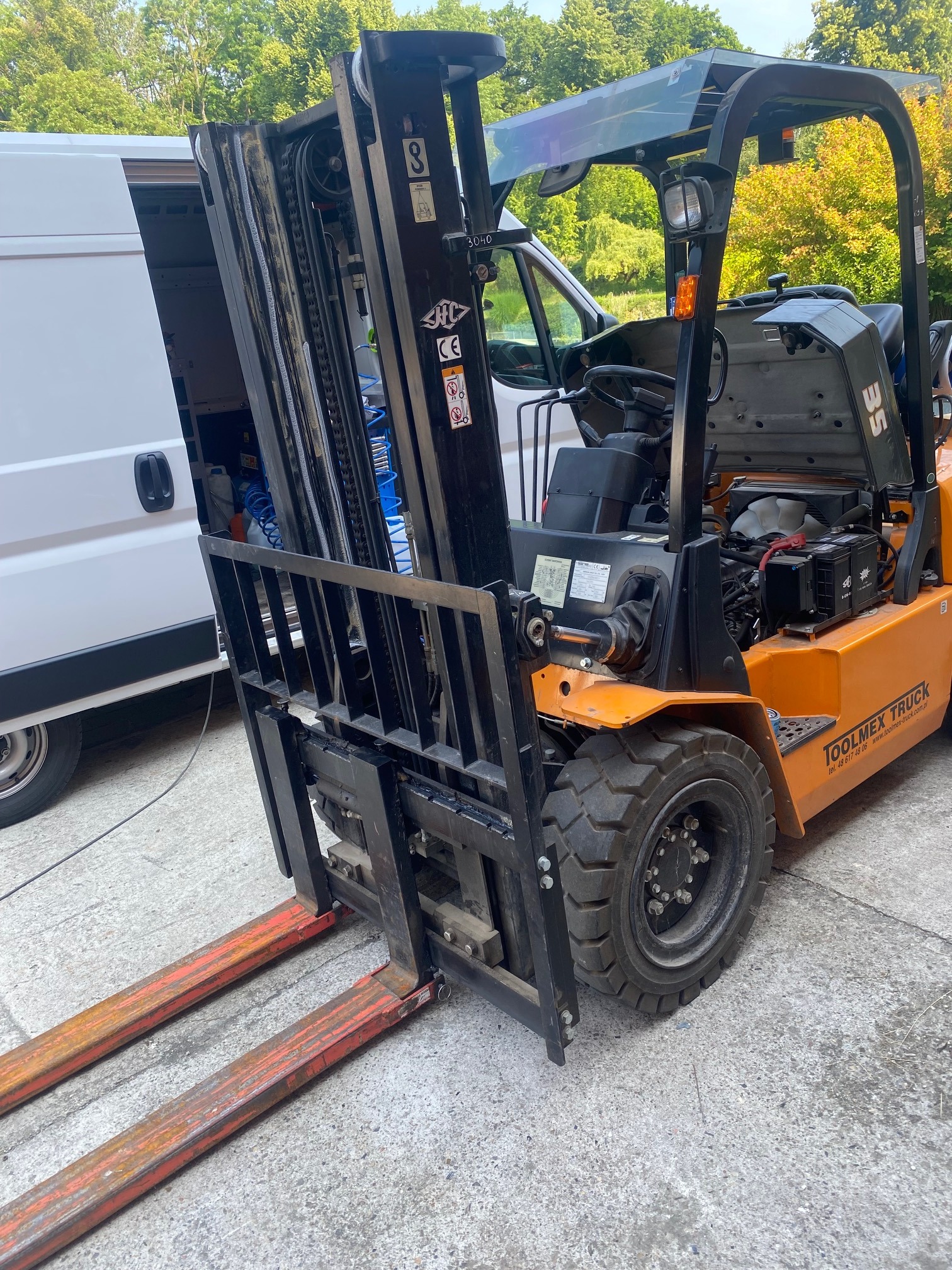 Pomarańczowy wózek widłowy marki Toolmex Truck z otwartą maską silnika, stojący obok białego busa z otwartymi drzwiami, prawdopodobnie podczas serwisu.