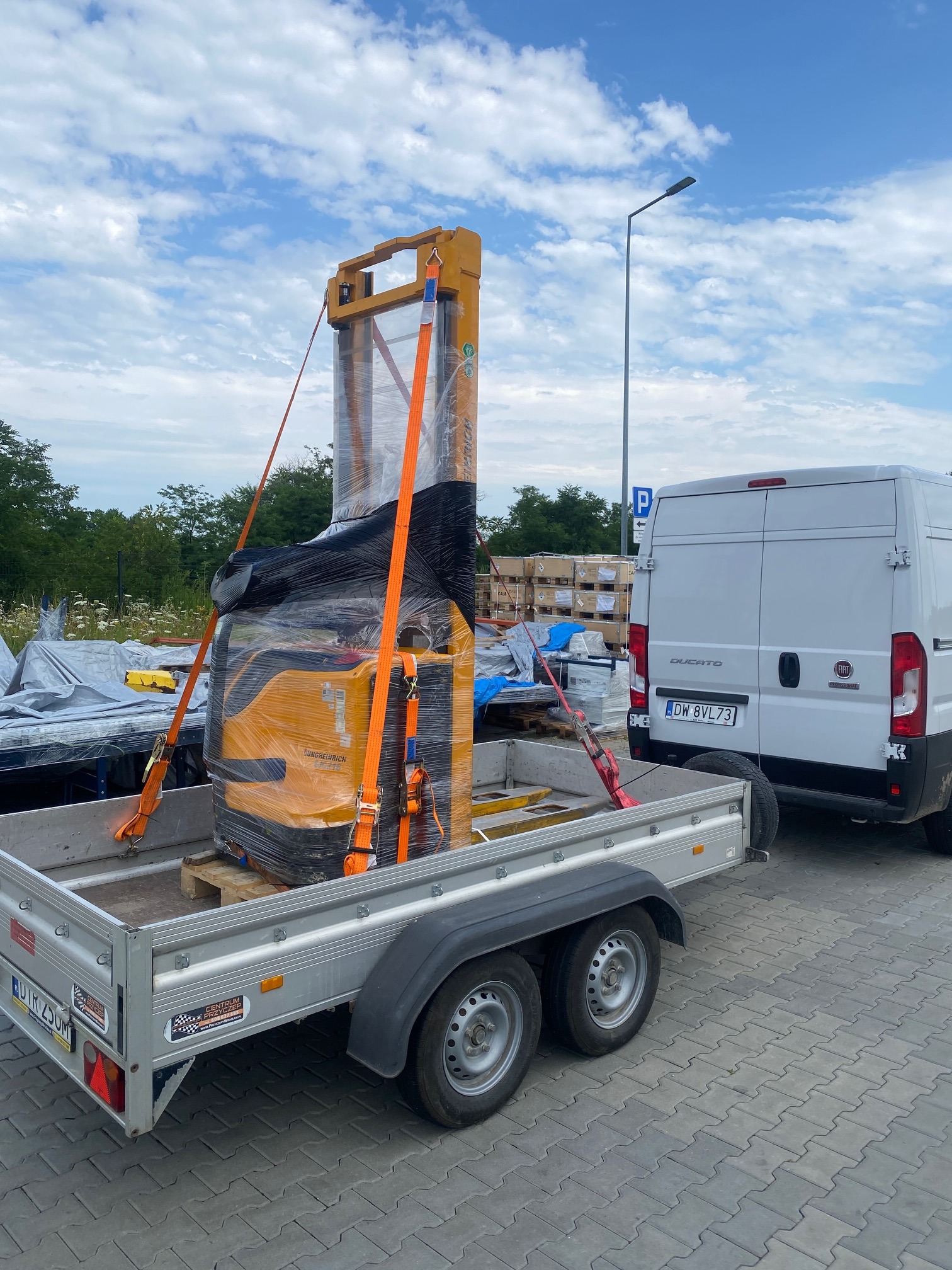 Nowy wózek widłowy Jungheinrich, owinięty folią i zabezpieczony pomarańczowymi pasami transportowymi, umieszczony na przyczepie samochodowej, gotowy do transportu, z widocznym fragmentem białego...