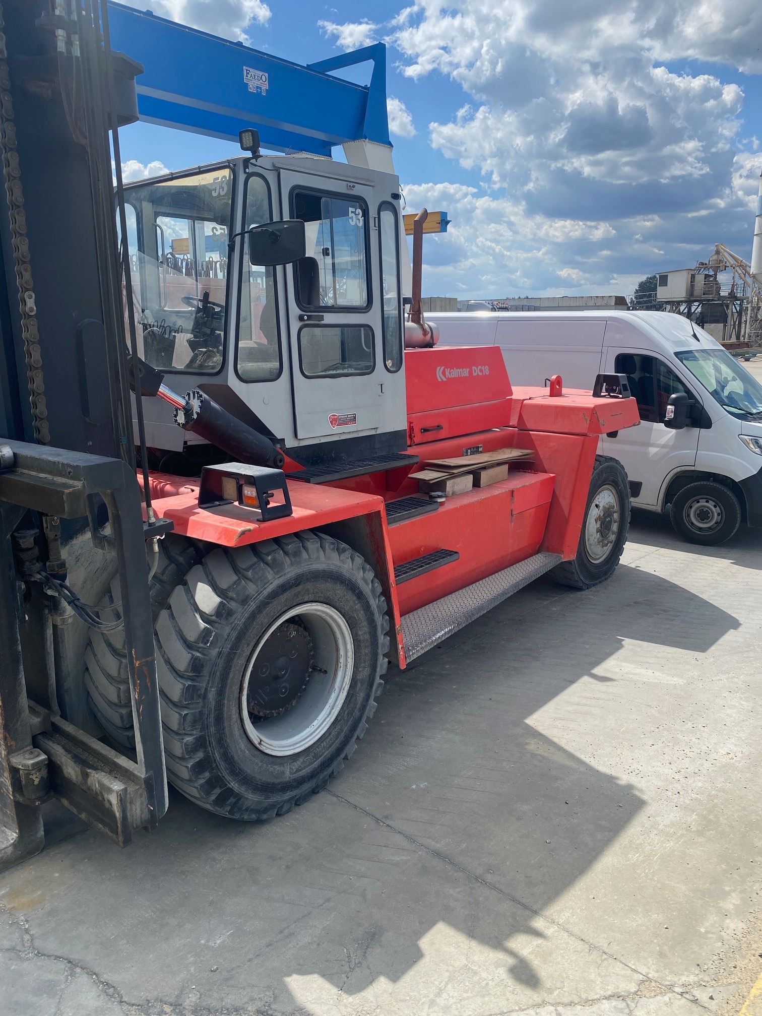 Czerwono-biały wózek widłowy Kalmar DC18 na betonowym placu, z widoczną kabiną operatora i masywnymi oponami, na tle białego samochodu dostawczego i częściowo zachmurzonego nieba.