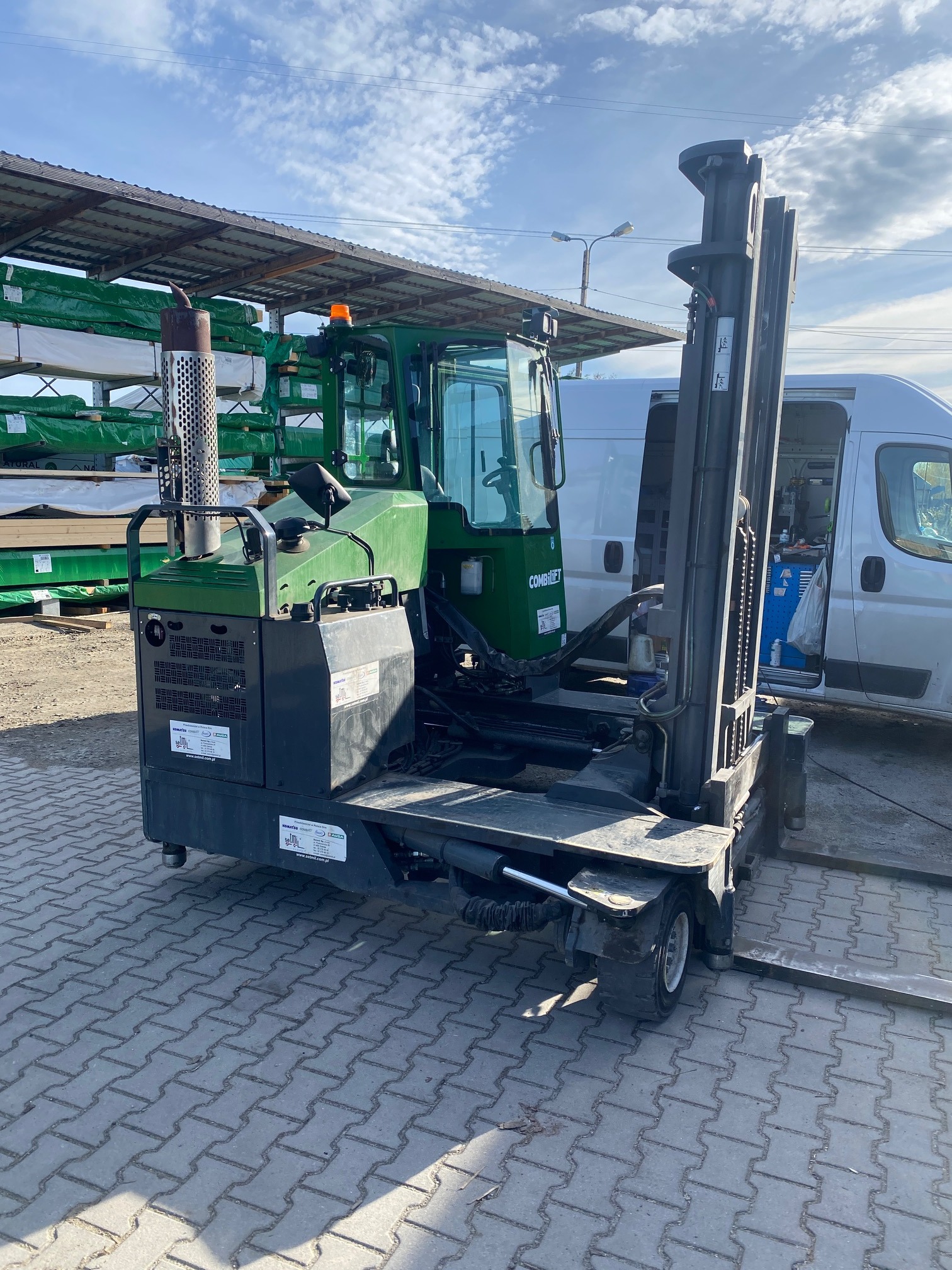 Wielokierunkowy wózek widłowy Combilift na zewnątrz, z widocznym kominem i otwartym busem w tle, na brukowanej powierzchni.