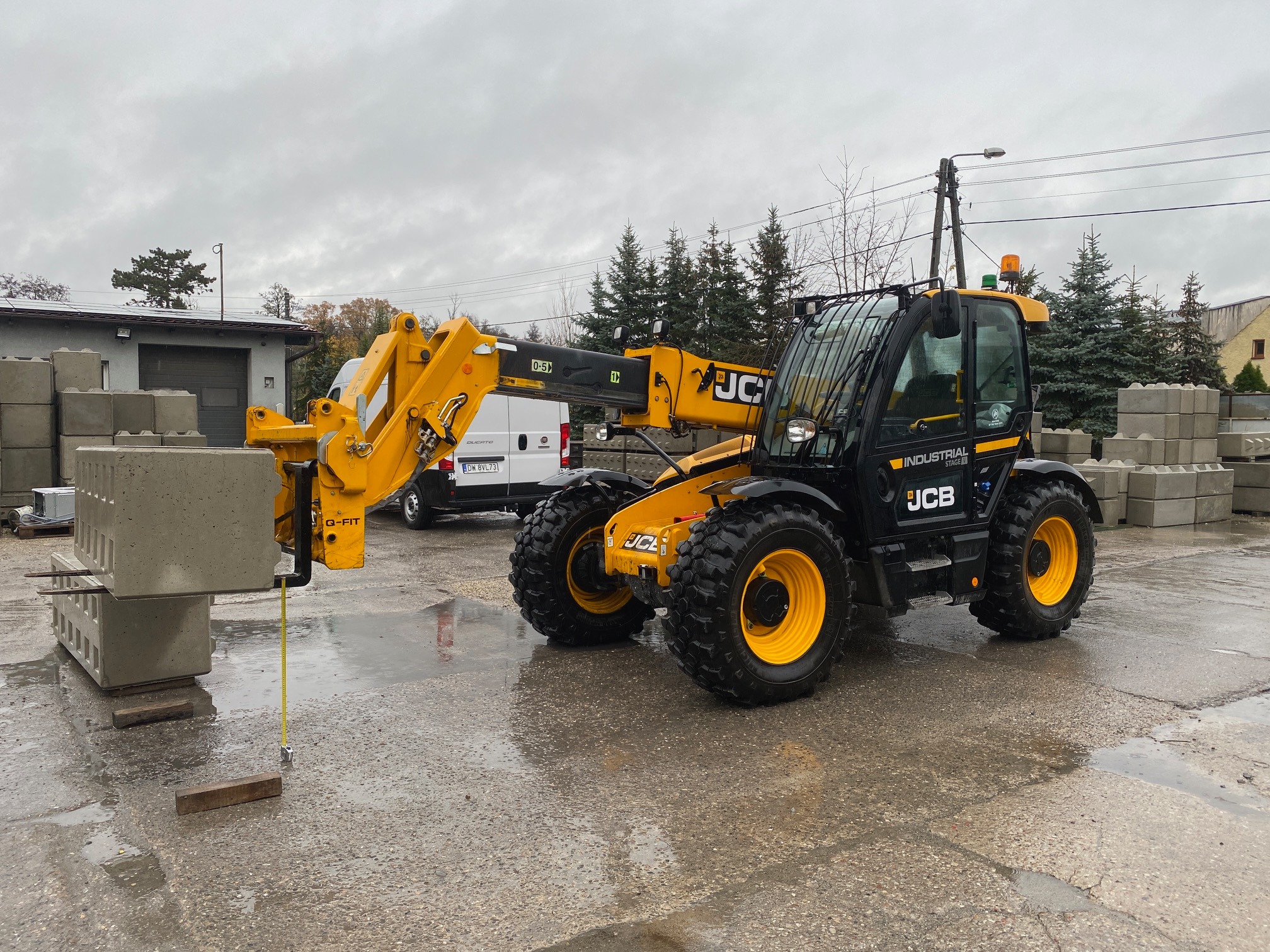 Żółto-czarny wózek widłowy teleskopowy JCB podnosi betonowy element konstrukcyjny na mokrym placu budowy, w tle budynek i zaparkowany biały samochód dostawczy.
