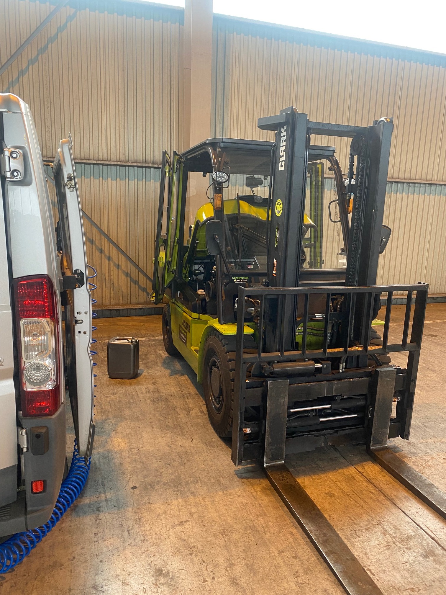 Żółto-czarny wózek widłowy marki Clark stoi w hali obok otwartego samochodu dostawczego, widoczny kanister, podłoga betonowa.