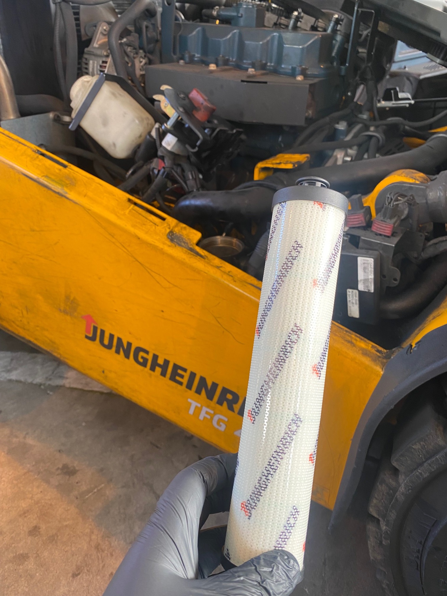 Filtr oleju silnikowego w rękawicy na tle otwartej maski wózka widłowego Jungheinrich TFG 4 z widocznym silnikiem.